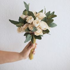 a person holding a bouquet of white roses and greenery in their hand, against a white wall