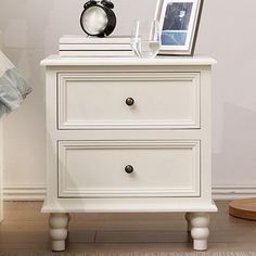 a night stand with two drawers and a clock on top, next to a bed