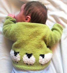 a baby in a green sweater laying on top of a white bed next to a pillow