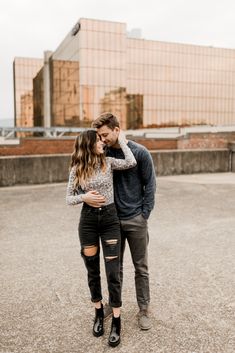 a man and woman standing next to each other in an empty parking lot with tall buildings behind them