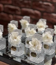 small clear vases with white flowers are arranged on a glass tray in front of a brick wall