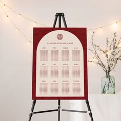 a red and white wedding seating chart on a easel with lights in the background