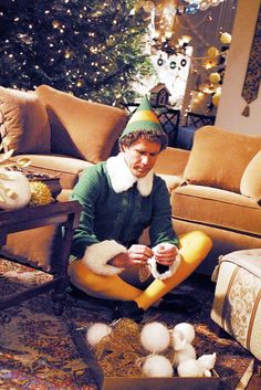 a man sitting on the floor in front of a christmas tree wearing a gnome hat