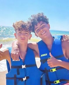 two boys in life vests on the beach giving thumbs up