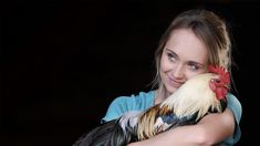 a girl holding a rooster in her arms and looking at the camera with an intense look on her face