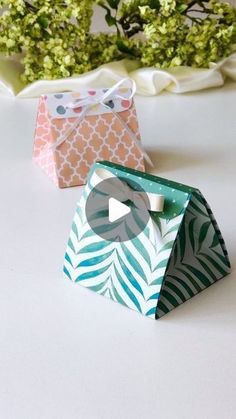 two small gift bags sitting next to each other on top of a white table with flowers in the background