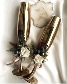 two gold vases with white flowers and greenery in them on a white sheet