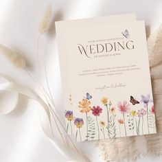 a wedding card with watercolor flowers and butterflies on it next to some white feathers