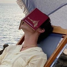 a woman laying on top of a beach chair next to the ocean with a book on her head