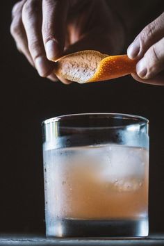 a person holding an orange peel over a glass of water with ice and liquid in it