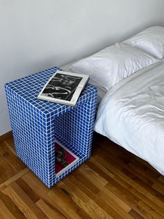 a small blue table with a magazine on it next to a white bed and wooden floor