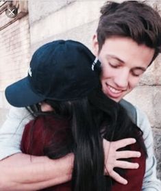 a man and woman hugging each other in front of a stone wall with a baseball cap on his head