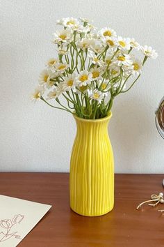 a yellow vase filled with white daisies on top of a wooden table next to an alarm clock