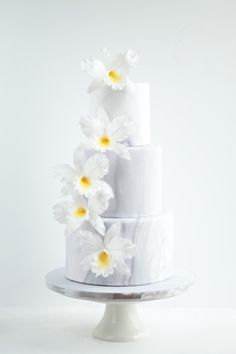 a three tiered cake with white and yellow flowers on the top, in front of a white background