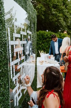 a woman writing on a wall with people in the background at an outdoor event, surrounded by greenery