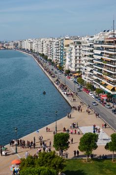 people are walking along the water in front of some buildings and cars on the street