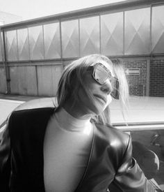 a woman with glasses leaning on a car in front of a building and looking off into the distance