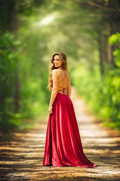 a woman in a red dress is standing on a dirt road with trees behind her