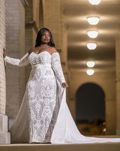 a woman in a white dress standing next to a brick wall wearing a long sleeved gown