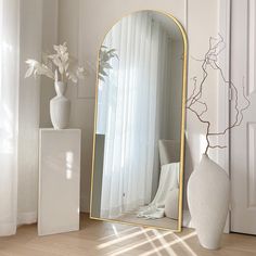 a white vase sitting next to a large mirror on top of a wooden floor in front of a window