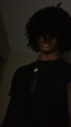 a young man with curly hair wearing a black t - shirt and silver necklace, standing in the dark