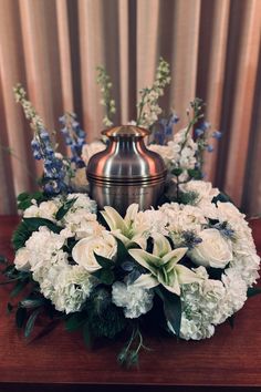 white flowers and greenery are arranged in a metal container on a wooden table next to a curtain