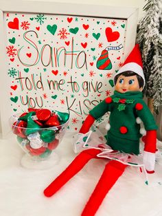 an elf doll sitting next to a bowl of candy and a christmas card with santa's help me give written on it