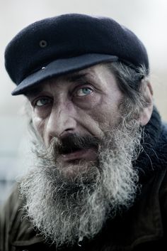 an old man with a long beard wearing a hat and looking at the camera while standing outside