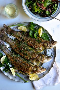 grilled fish with herbs and lemons on a plate next to a bowl of salad