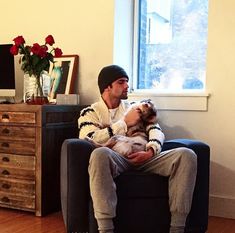 a man sitting on a couch holding a dog in his lap and looking out the window