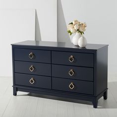 a black dresser with gold handles and flowers on top, against a white wall in an empty room