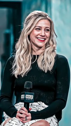 a woman sitting down with a microphone in her hand and smiling at the camera while talking to someone