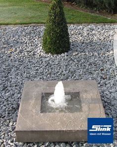 a stone fountain in the middle of a graveled area with trees and bushes behind it