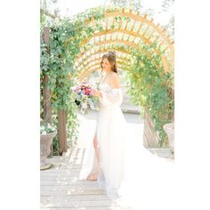 a woman in a long white dress is standing under an arch with flowers and greenery