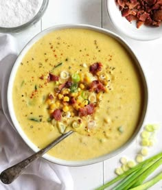 a bowl of corn chowee with bacon and green onions next to it on a white table