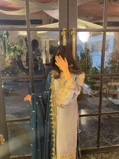 a woman standing in front of a glass door wearing a white and blue outfit with gold embroidery on the sleeves