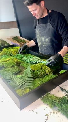 a man in an apron and gloves working on a moss covered table with green plants