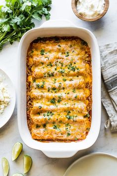 an enchilada casserole in a white dish with limes and cilantro on the side