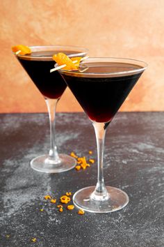 two martini glasses with orange peel garnish on the rim and one filled with black liquid