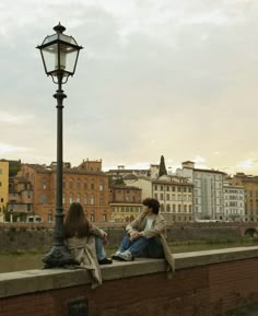 two women sitting on a wall next to a street light and lamp post with buildings in the background