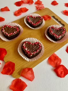 three heart shaped cookies sitting on top of a wooden board next to red petals and rose petals