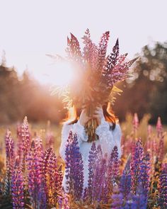 a woman standing in the middle of a field with purple flowers on her head and sun shining behind her