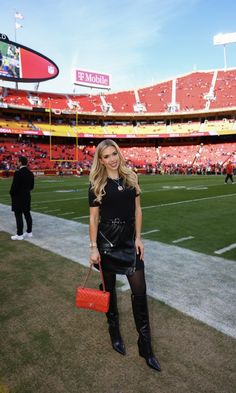 a woman standing in the middle of a football field wearing black boots and holding a red purse