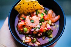 a hand holding a small bowl filled with shrimp and veggies next to a tortilla chip