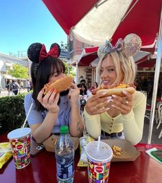 two women sitting at a table eating hot dogs and drinking sodas with mickey ears on their heads