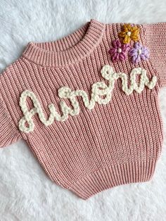 a pink sweater with flowers on it and the word baby written in white letters is laying on a fluffy surface