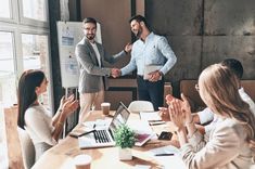 two men shaking hands over a table with other people in business attire at the same time