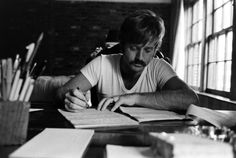 a man sitting at a table writing on paper with pencils in front of him