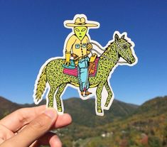 a hand holding up a sticker depicting a man riding a horse in the mountains