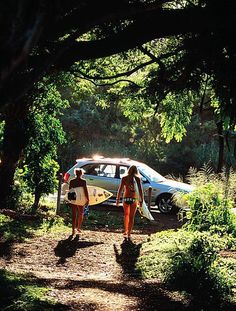 two women walking down a dirt road with surfboards in their hands and a car parked behind them